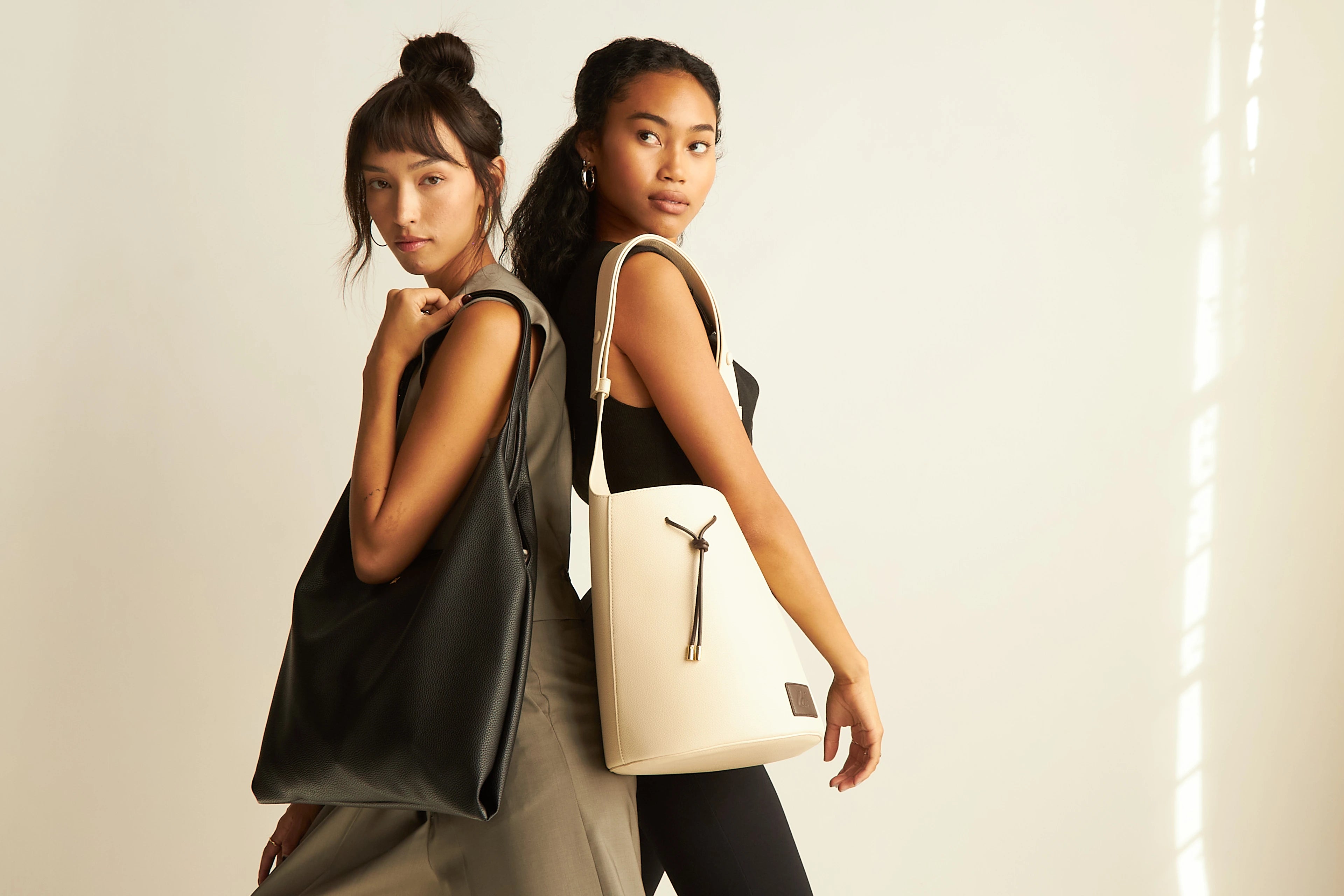 Two models standing together, one holding a black tote bag and the other holding a white bucket tote bag.